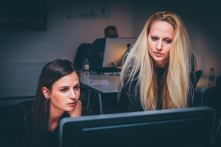 Biuro. Dwie kobiety – jedna stoi, druga siedzi – patrzą na monitor komputera