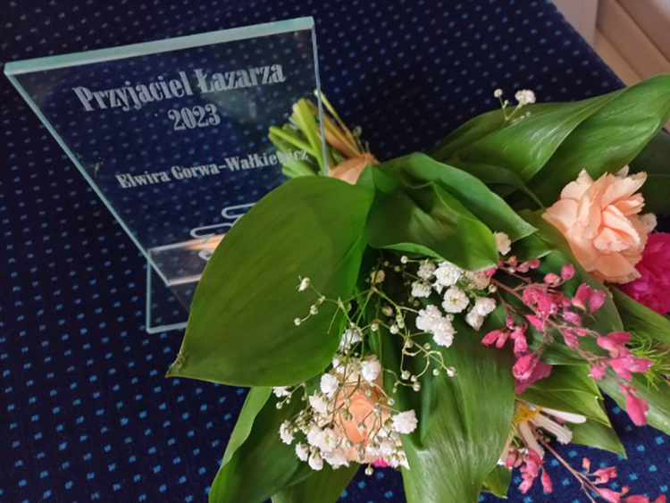Na stole leży statuetka z napisem "Przyjaciel Łazarza. Elwira Gorwa-Wałkiewicz" i bukiet kwiatów 