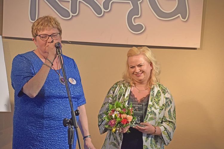 Jedna kobieta stoi przed mikrofonem. Obok niej kobieta z kwiatami