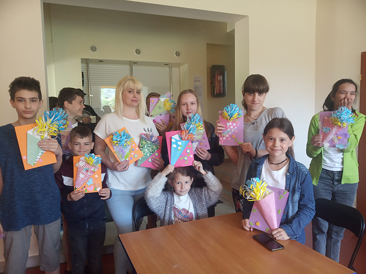 Kobieta stoi z dziećmi, pokazują papierowe kwiaty i laurki