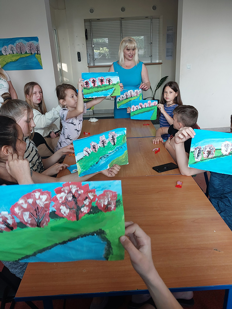 Kobieta stoi, dzieci siedzą, pokazują rysunki