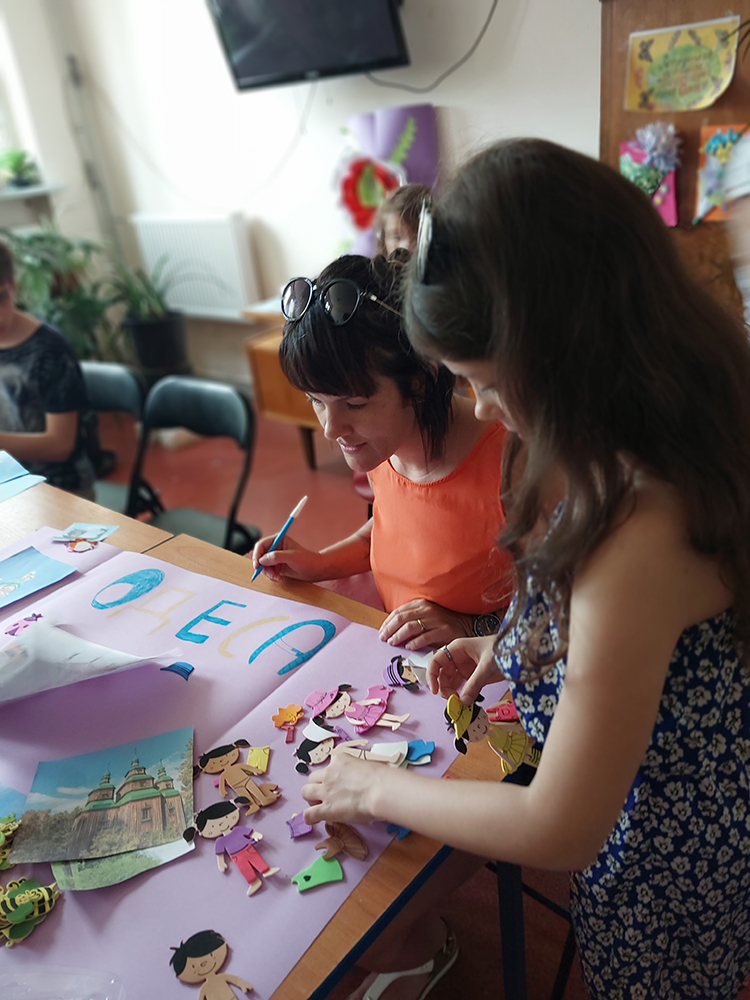 Kobieta trzyma pisak, obok dziewczynka wybiera papierowe figurki